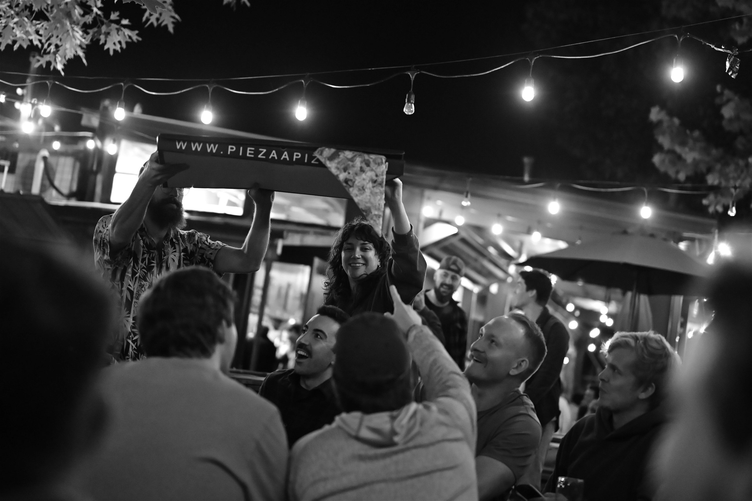 A black and white photo captures a lively scene at an outdoor gathering under string lights. A person in the center is holding up a large pizza box from "Piezaa Pizza" with a slice of pizza in hand, while others around them look up, smiling and pointing at the pizza. The ambiance suggests a casual, festive atmosphere with people enjoying food and company.