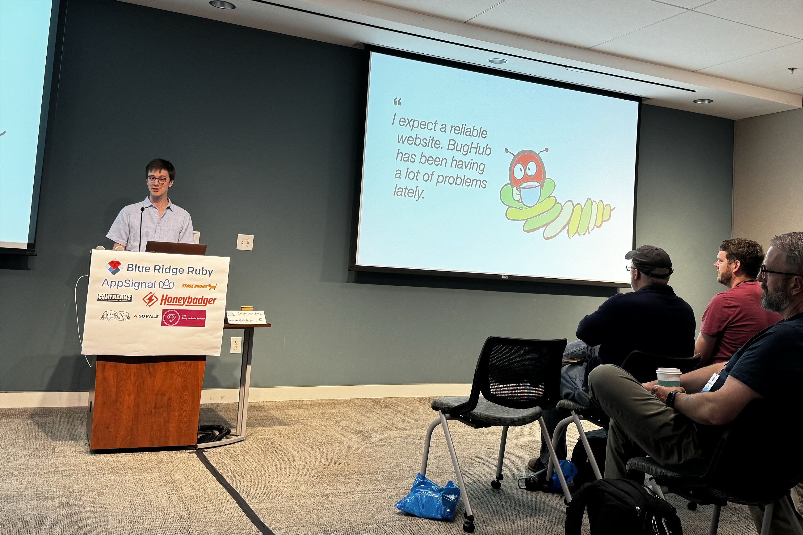 Daniel Colson at a Blue Ridge Ruby conference presenting a slide titled "I expect a reliable website. BugHub has been having a lot of problems lately." with a cartoon bug image, in front of an audience of attentive attendees.