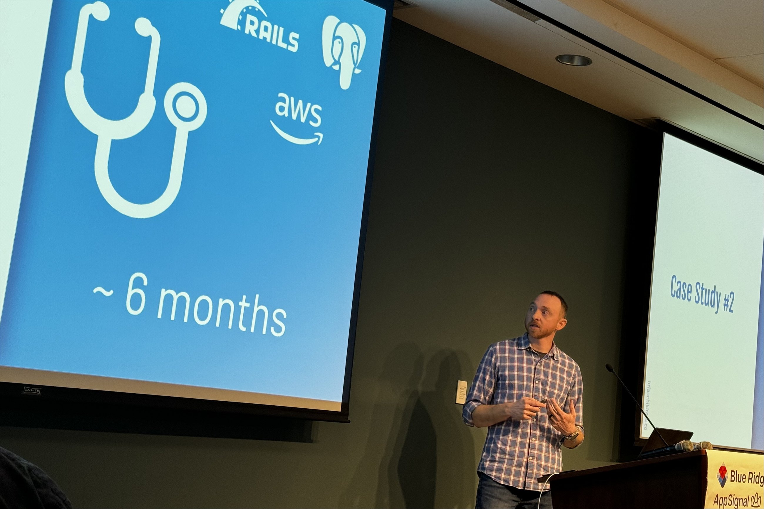 Brian Childress at Blue Ridge Ruby conference discussing a case study related to Rails and AWS, indicated by icons on a slide that reads "~ 6 months", standing beside a podium with a Blue Ridge Ruby conference banner.
