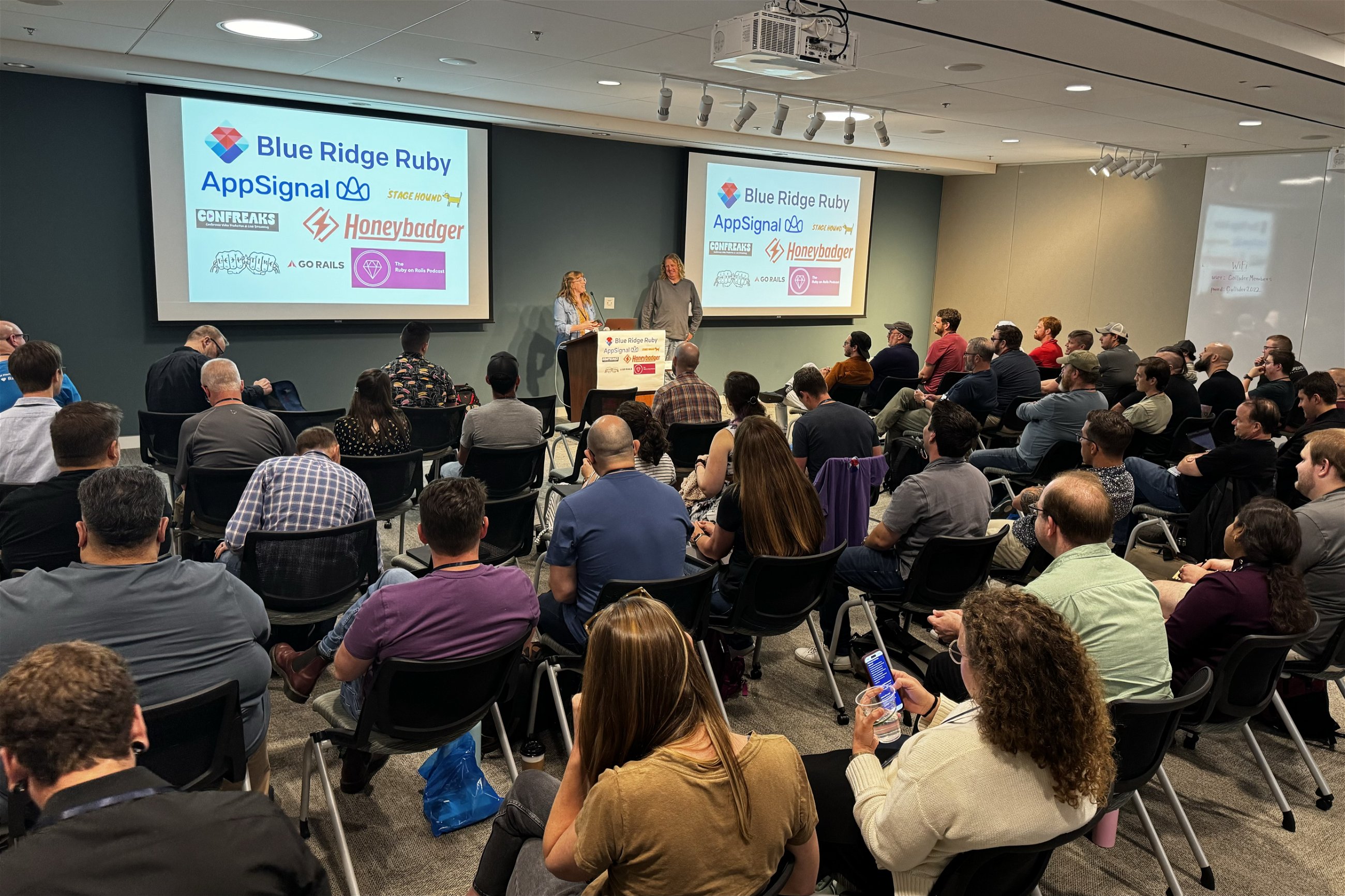 Two speakers addressing a large audience at the Blue Ridge Ruby conference, with sponsor banners including Honeybadger visible behind them, in a room filled with seated attendees listening attentively.