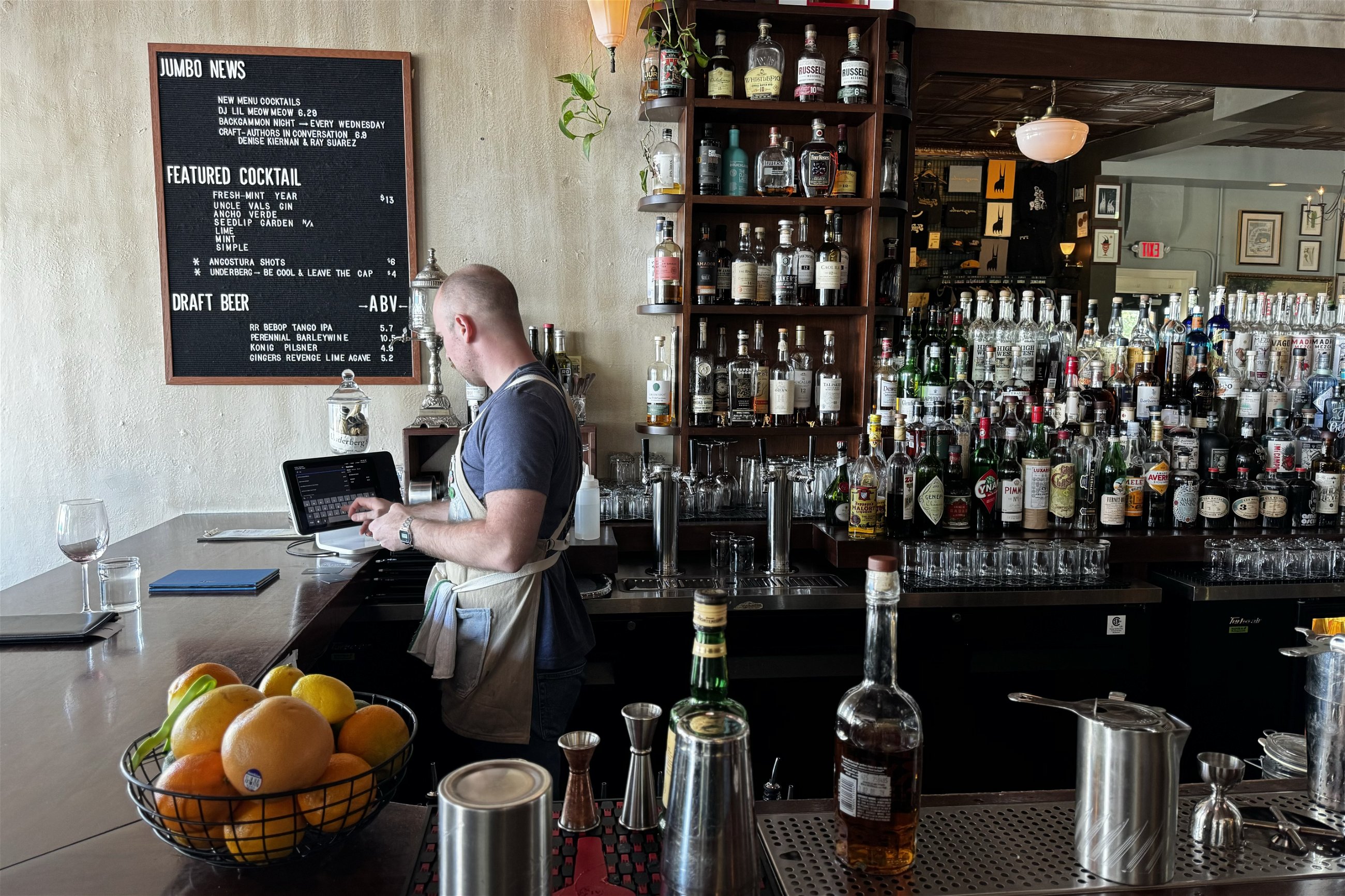 Man working on an iPad at a bar counter, surrounded by an extensive selection of spirits displayed on shelves, with a blackboard listing featured cocktails and draft beers above him.