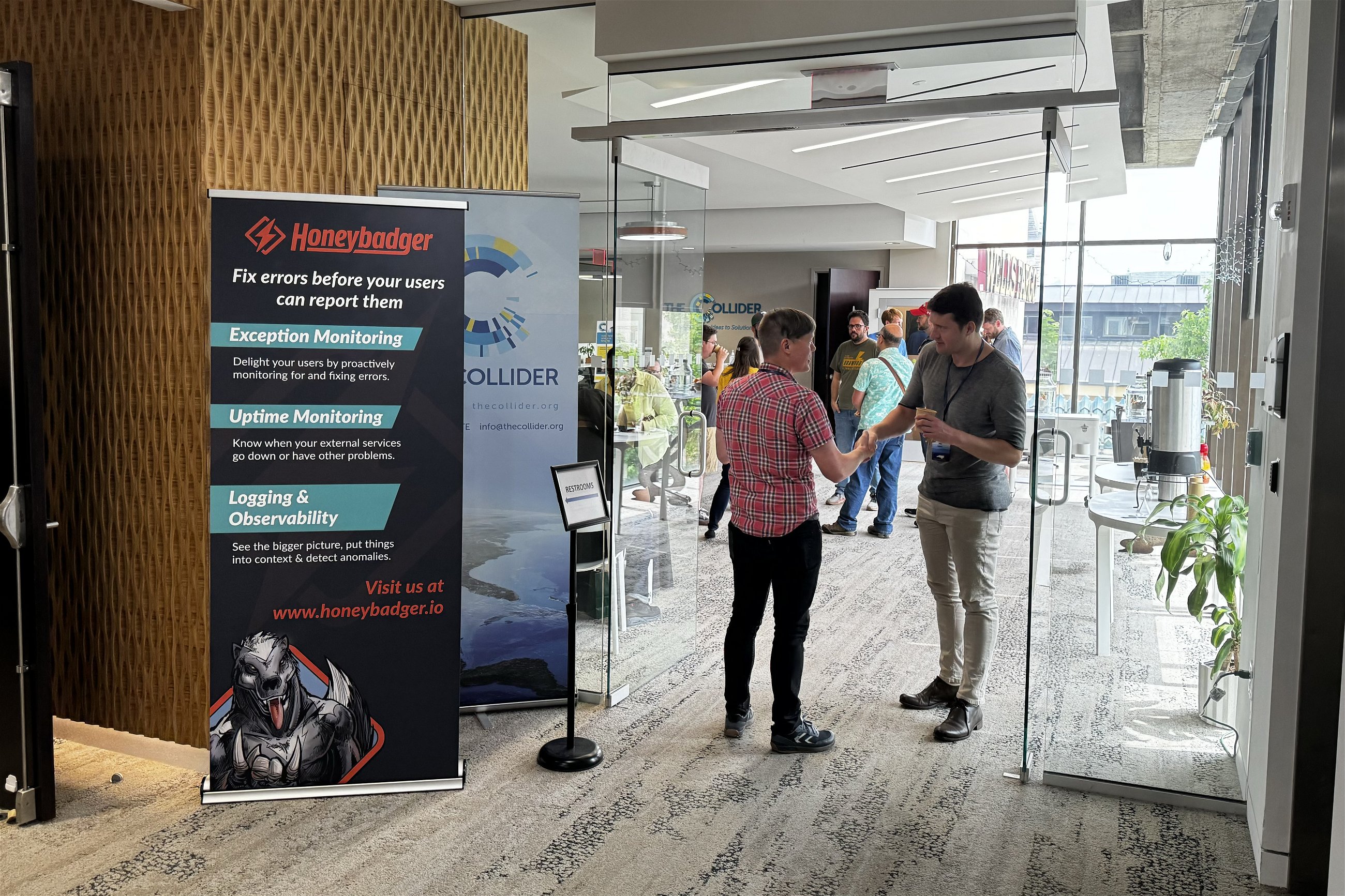 Two people shaking hands in a conference setting near a Honeybadger promotional banner, with other attendees visible in the background through glass doors. The banner highlights services like Exception Monitoring, Uptime Monitoring, and Logging & Observability.