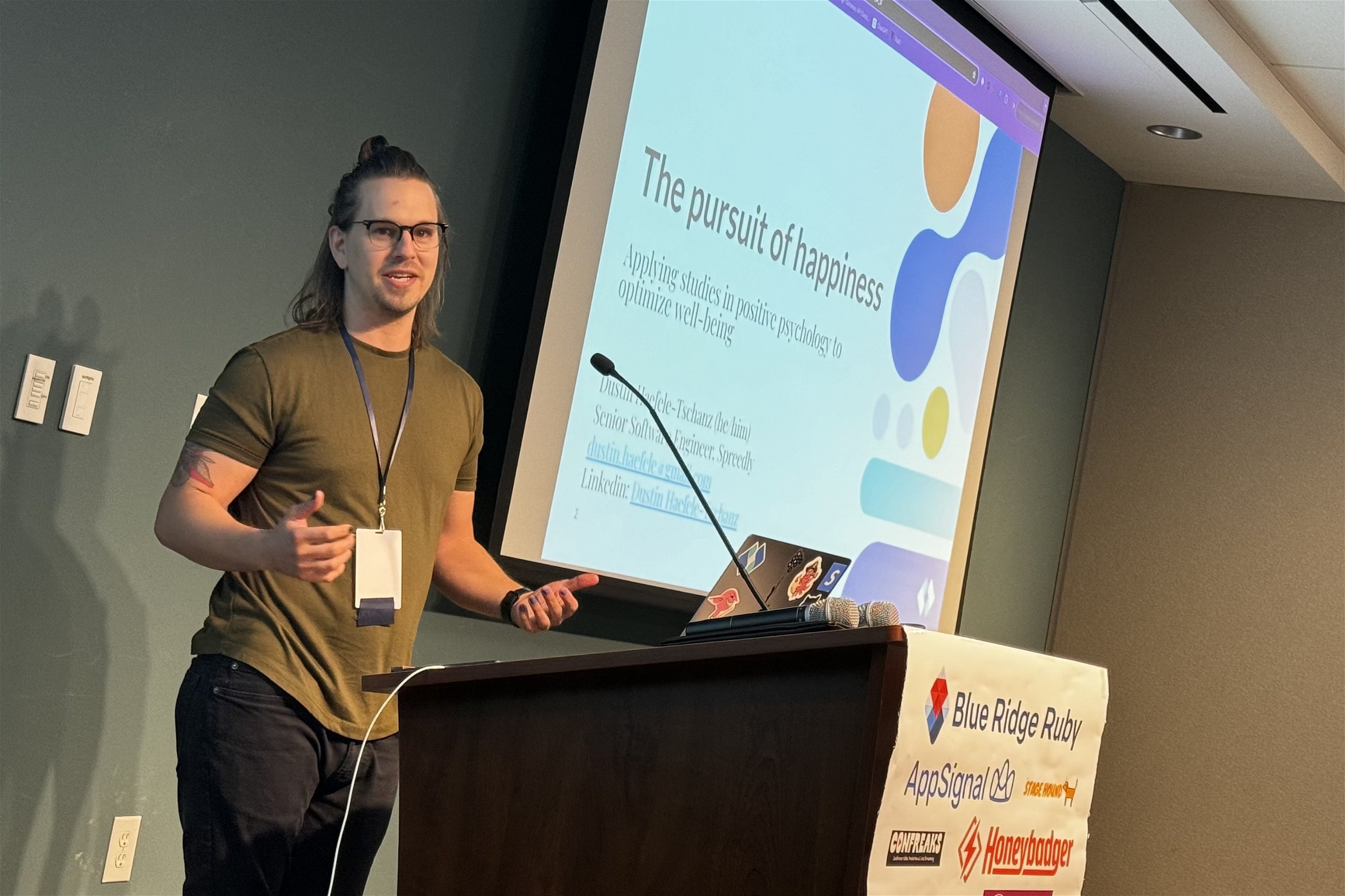Dustin Haefele-Tschanz presents "The Pursuit of Happiness: Applying Studies in Positive Psychology to Optimize Well-being" at Blue Ridge Ruby conference, engaging the audience with a slide displayed behind him, featuring his contact information and professional title.
